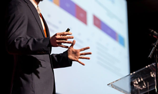 Presentation Camera Rentals A man giving a presentation at a podium with a large projection screen displaying information behind him