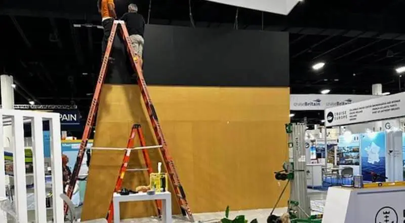 Video Wall Rentals for Trade Shows A blank wall at a trade show with work equipment around it. A man stands on top of a ladder working on some wiring.
